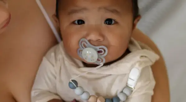 A baby with a pacifier, possibly experiencing pacifier nipple confusion, looks into the camera while being held by a parent.