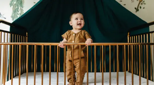 A happy baby stands in a golden crib, holding the bars. What do you need for a crib? A safe space, a soft mattress, and a smiling baby!