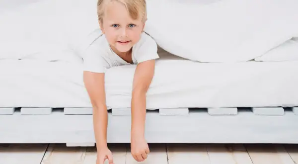 A young boy peeks out from under white sheets, his arms dangling. He looks ready for a new adventure, perhaps signaling it's time to transition to a floor bed.