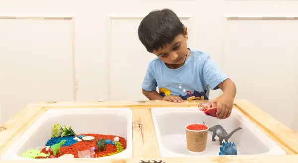 A boy pours red liquid into a sensory table with dinosaurs and colorful rice, exploring fun sensory table ideas.