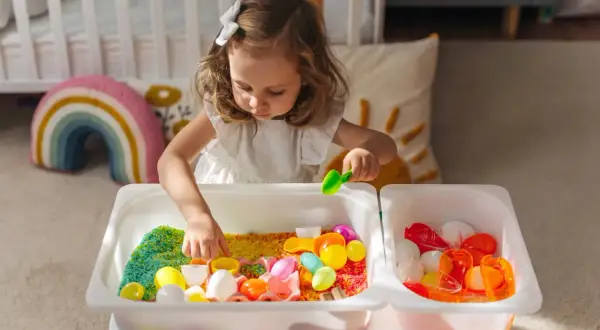 A young girl explores colorful rice and toys in sensory bins, demonstrating why sensory bins are important for early childhood development and learning through play.