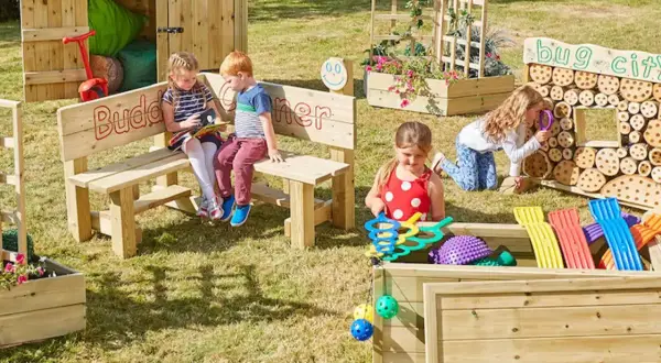 Children explore a vibrant Montessori garden, discovering nature's wonders. A bug city invites curiosity, while a planter box bursts with colorful blooms.