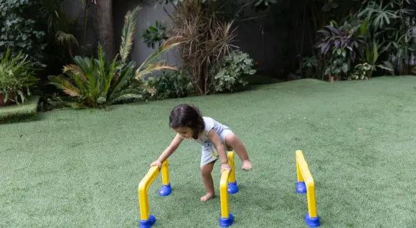 A toddler practices gross motor outdoor activities, balancing on yellow and blue hurdles in a grassy yard.