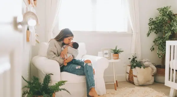 A mother cradles her baby in a cozy nursery, surrounded by soft decor and plants, hinting at peaceful nursery themes.