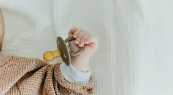 A tiny hand holds a pacifier that appears too big for a newborn, nestled in a soft, textured blanket.