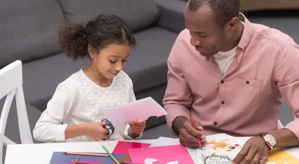 A father and daughter are crafting Mother's Day cards. The toddler cutting paper with scissors is focused on her task.