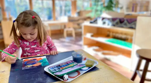 A young girl with pigtails paints at a table, exploring her creativity with watercolors. This scene captures the essence of Montessori nature-inspired learning.