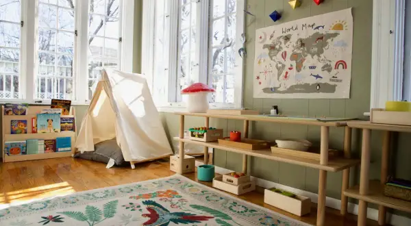 A bright, airy room with a cozy montessori home setup. A tent, world map, and wooden shelves filled with books and toys create a playful learning environment.