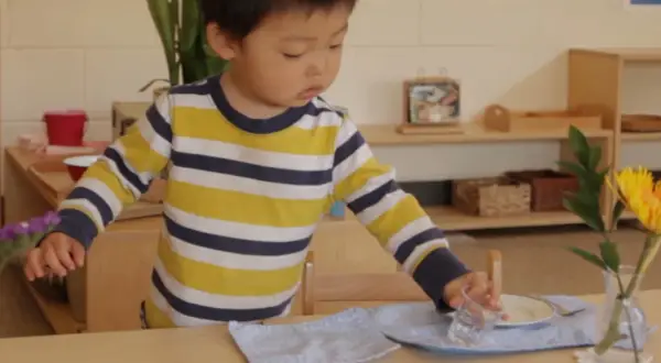 A young child, around 18 months old, practices setting a table in a Montessori environment, carefully placing a glass.