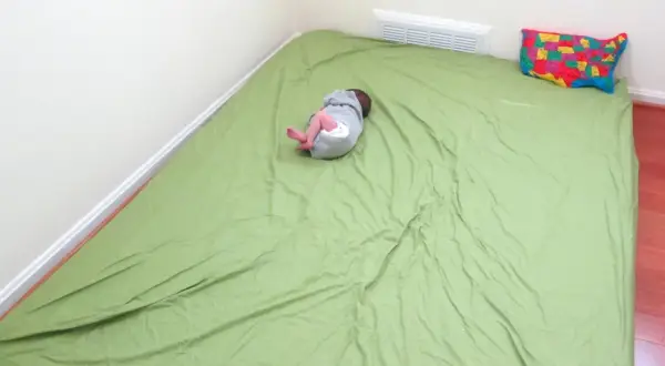 A baby sleeps on a green mattress on the floor, a simple setup for how to make a floor bed for baby. A colorful map pillow rests nearby.