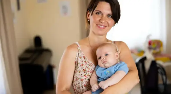 A smiling mother holds her 2-month-old baby, celebrating precious milestones. The baby wears a blue onesie with a yellow and blue heart.