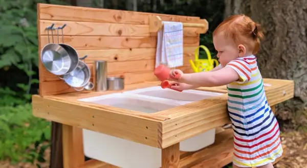 A toddler engages in Montessori movement activities, exploring a wooden mud kitchen with pots and a watering can.