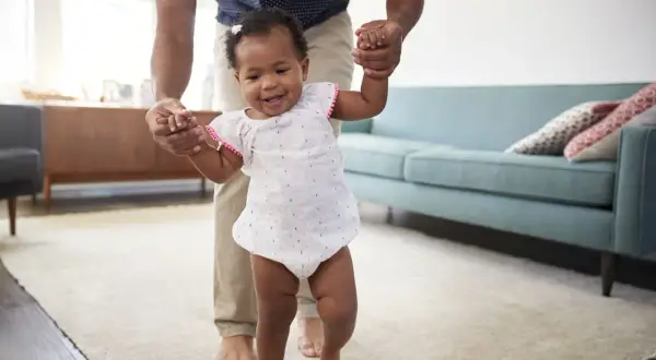 A father helps his baby practice gross motor activities for infants 12-18 months, taking first steps with a smile.