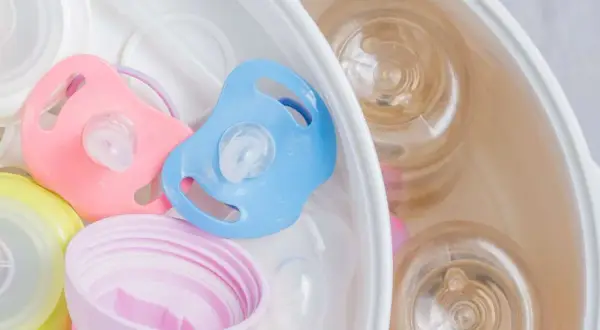 Pink and blue pacifiers sit in a sterilizer with baby bottles. Learn how to sterilize pacifier safely for your baby.