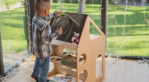 A young boy draws on a chalkboard easel, part of a Montessori backyard playhouse. He's erasing a drawing with a green eraser.