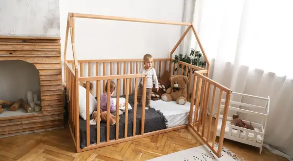 Two children play in a wooden playpen floor bed. One reads a book, the other stands, with a teddy bear nearby.