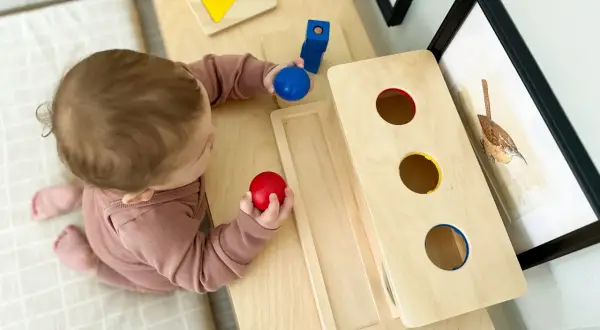A baby explores Montessori activities for 10 month old baby, holding red and blue balls near a wooden drop box and shape puzzle.