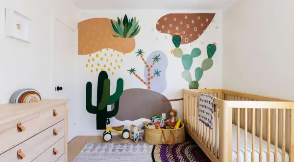 A colorful gender neutral nursery with a desert mural, wooden crib, dresser, and toys.