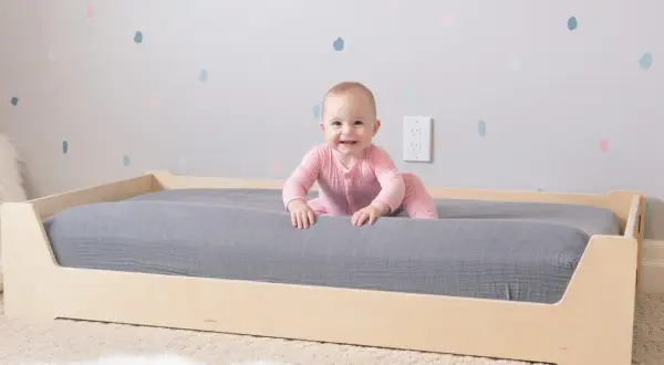 A smiling baby in pink pajamas explores a Montessori bed with a grey mattress. The wooden bed frame is low to the ground, perfect for little adventurers.