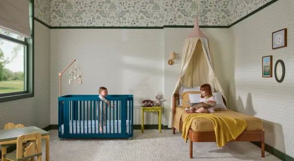 A teal crib offers bold crib color ideas in a nursery with whimsical wallpaper. Two children enjoy the room.