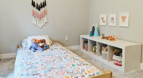 A cozy floor bed setup with a woodland-themed duvet cover, dolls, and a wall hanging. A white cube shelf holds books and toys.