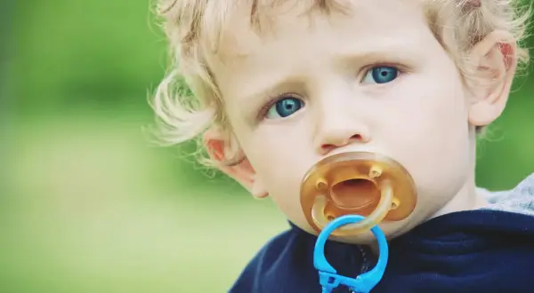 A toddler with blue eyes and blonde hair has a pacifier in their mouth. This image prompts the question: can pacifiers cause cavities?