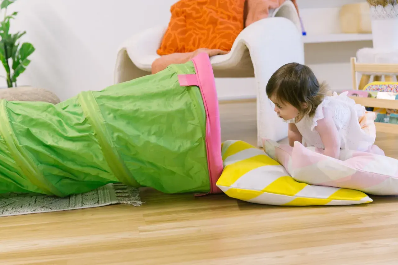 A toddler explores a green play tunnel, engaging in fun climbing activities. Colorful pillows add to the playful scene.
