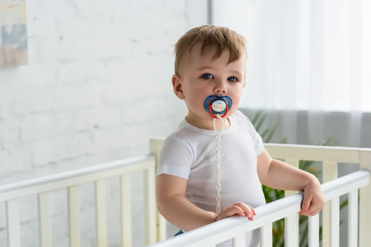 A baby in a crib with a pacifier. Some wonder if a pacifier can cause speech delay, but this little one seems content.