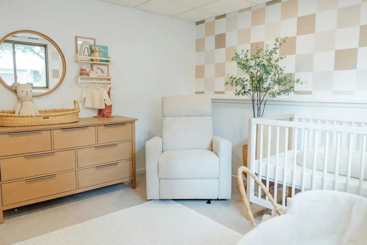 Cozy and modern nursery with a wooden dresser, plush recliner, and crib. This room offers sweet, cute baby room ideas with its neutral palette and playful checkered wallpaper.