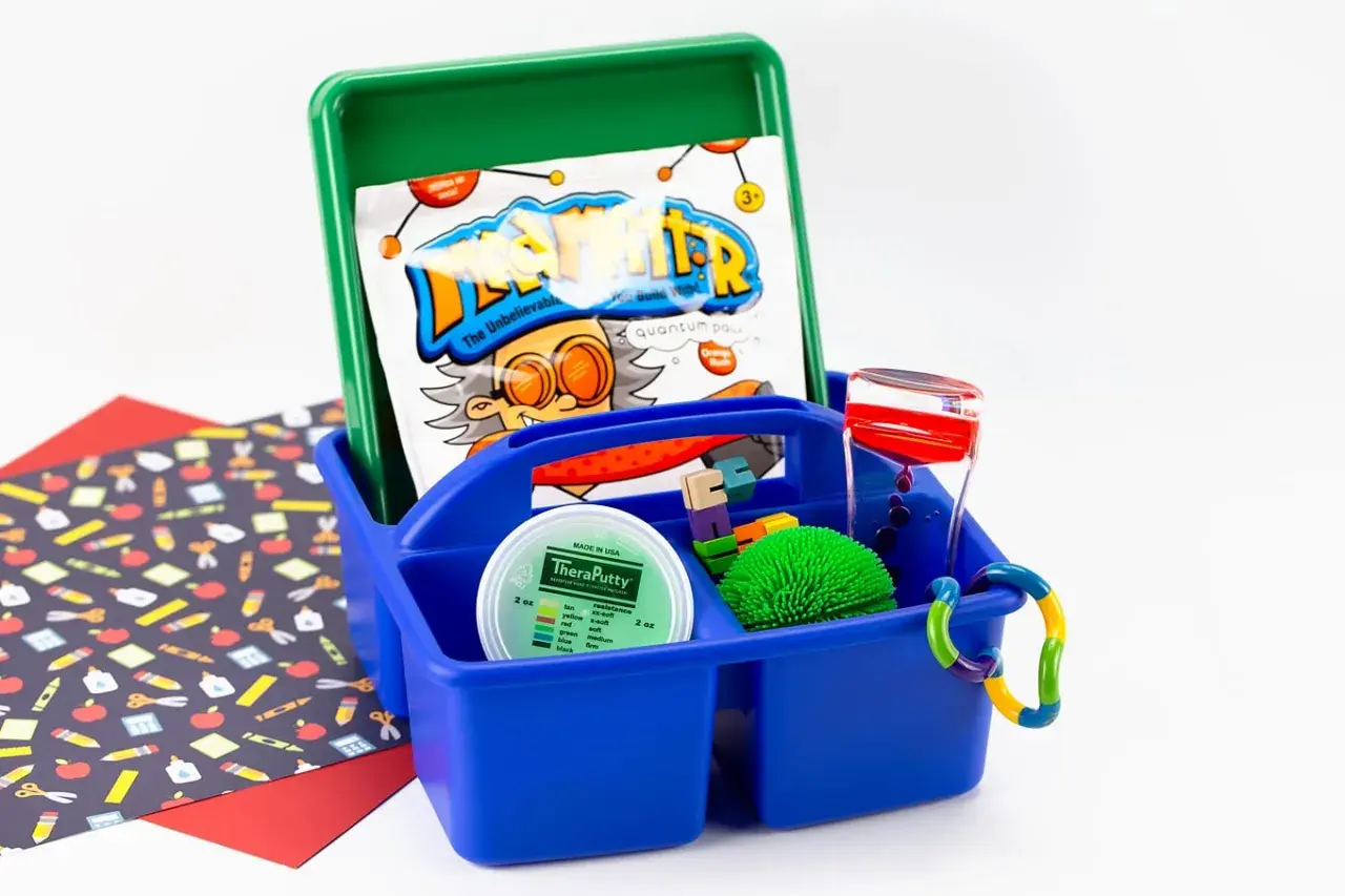 A blue caddy holds items for a sensory box: TheraPutty, a liquid timer, a spiky ball, and building blocks.