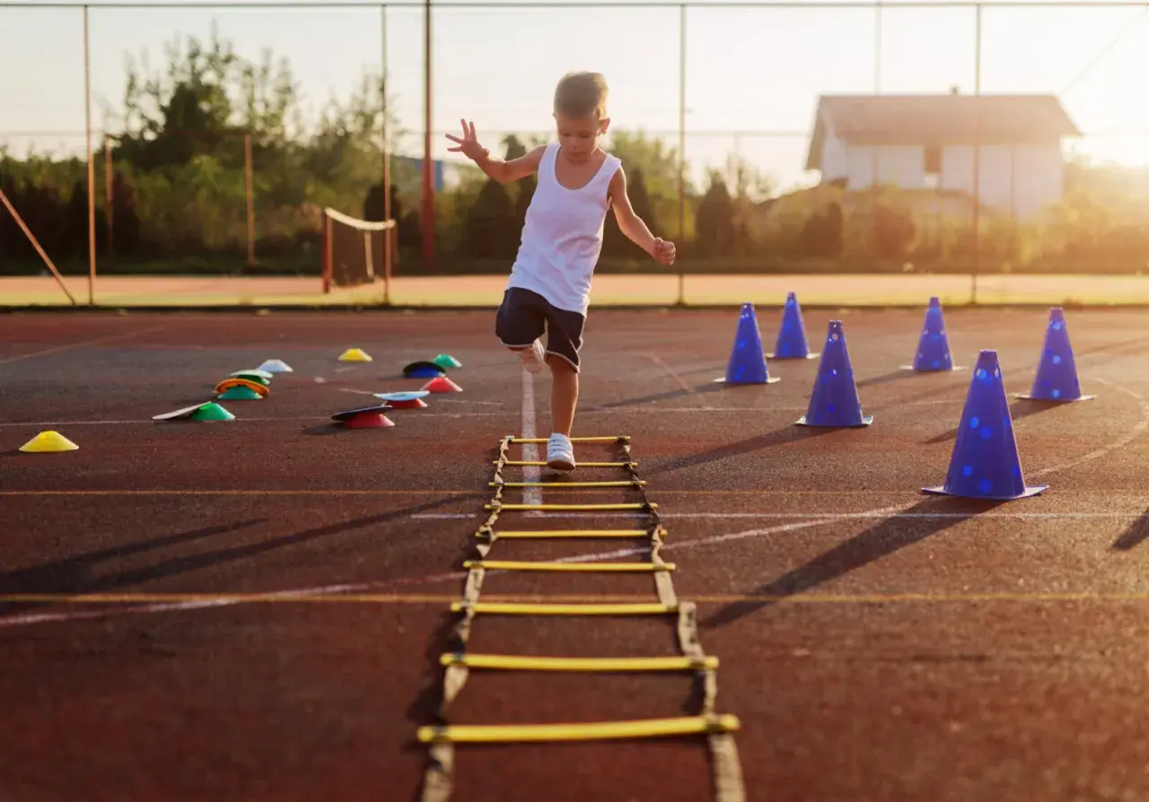 A young boy practices gross motor activities for 5 year olds, stepping through an agility ladder on a sports field with cones.