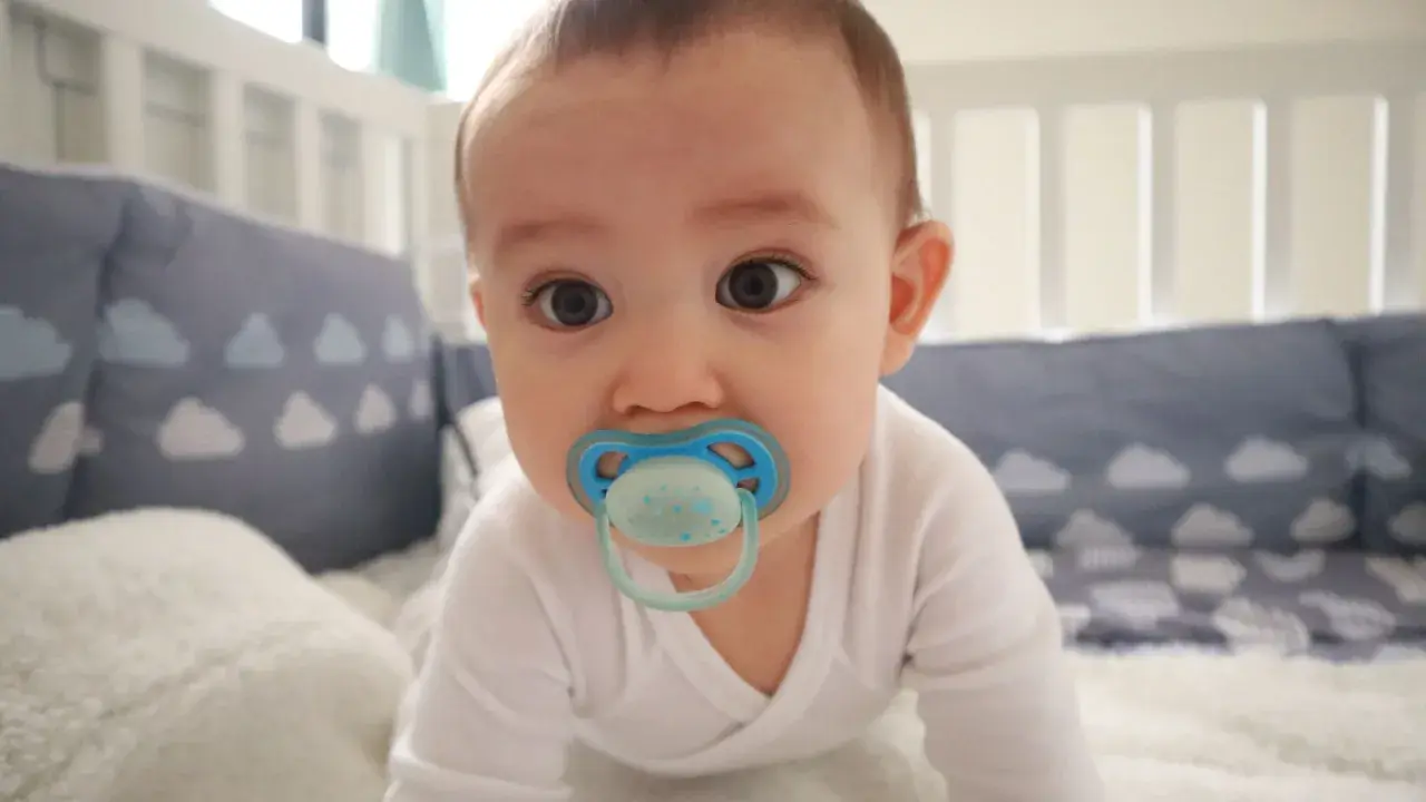 A baby with a blue orthodontic pacifier in a crib. Are orthodontic pacifiers better for developing smiles?