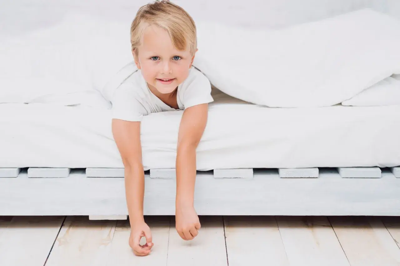 A young boy peeks out from under white sheets, his arms dangling. He looks ready for a new adventure, perhaps signaling it's time to transition to a floor bed.