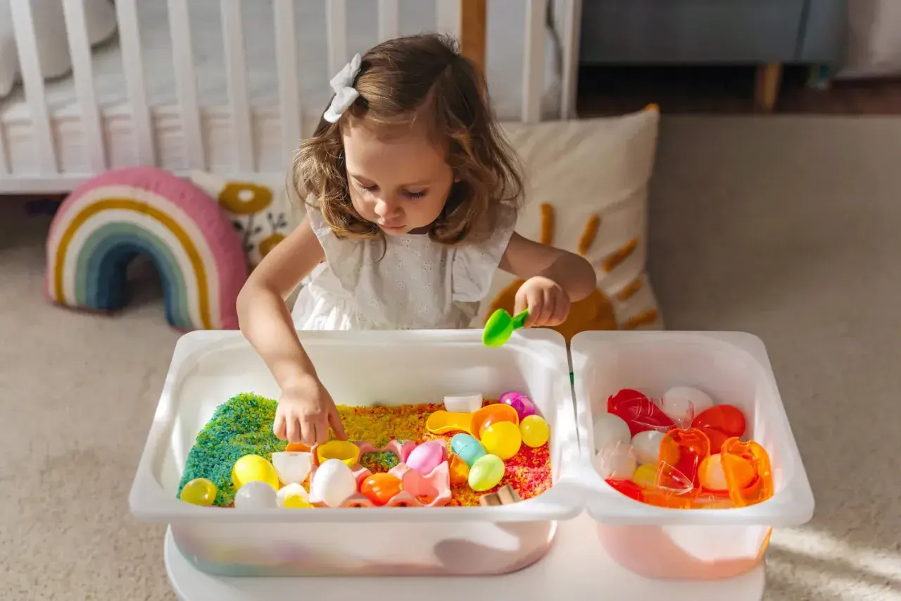 A young girl explores colorful rice and toys in sensory bins, demonstrating why sensory bins are important for early childhood development and learning through play.