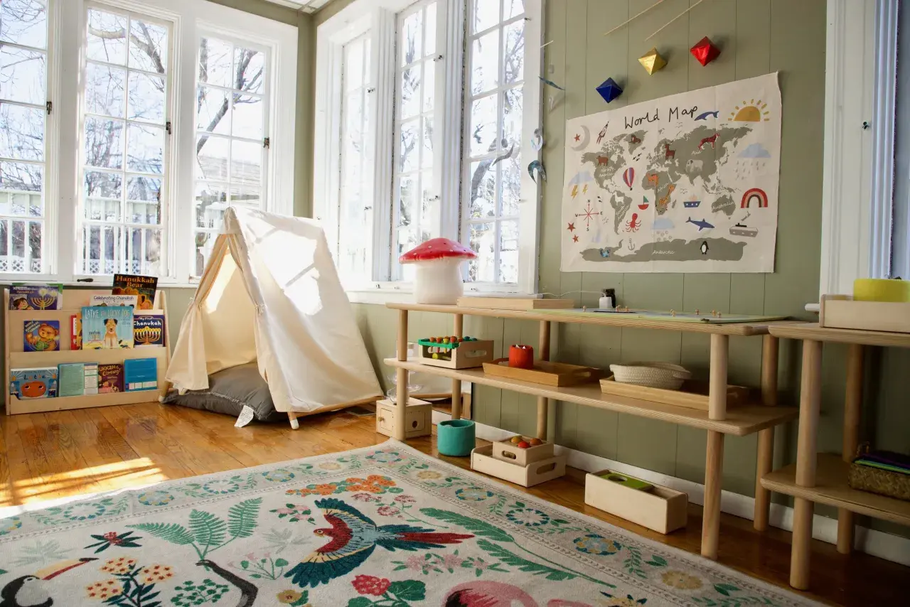 A bright, airy room with a cozy montessori home setup. A tent, world map, and wooden shelves filled with books and toys create a playful learning environment.