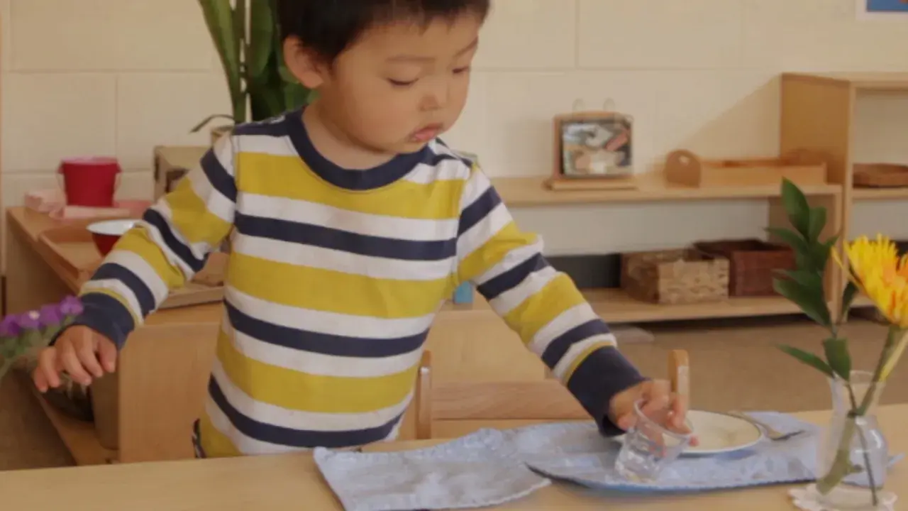 A young child, around 18 months old, practices setting a table in a Montessori environment, carefully placing a glass.