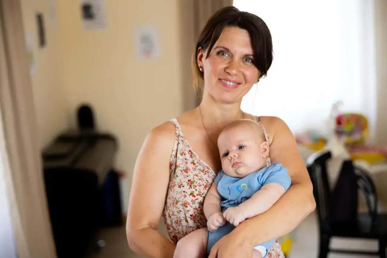 A smiling mother holds her 2-month-old baby, celebrating precious milestones. The baby wears a blue onesie with a yellow and blue heart.