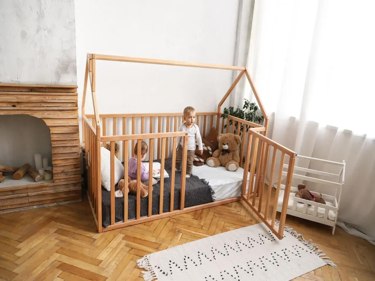 Two children play in a wooden playpen floor bed. One reads a book, the other stands, with a teddy bear nearby.