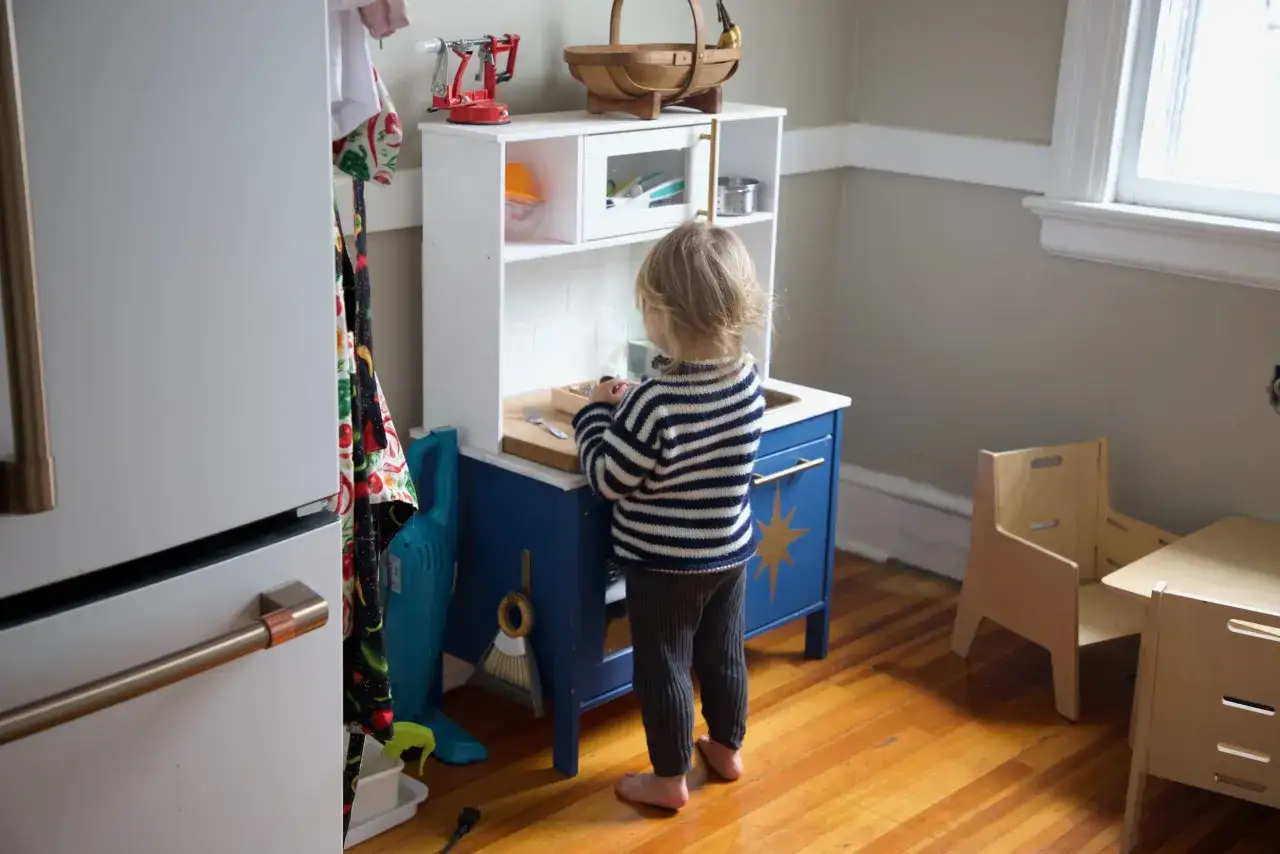 A child plays at a blue toy kitchen, part of a Montessori home setup. A wooden learning tower and table are nearby.