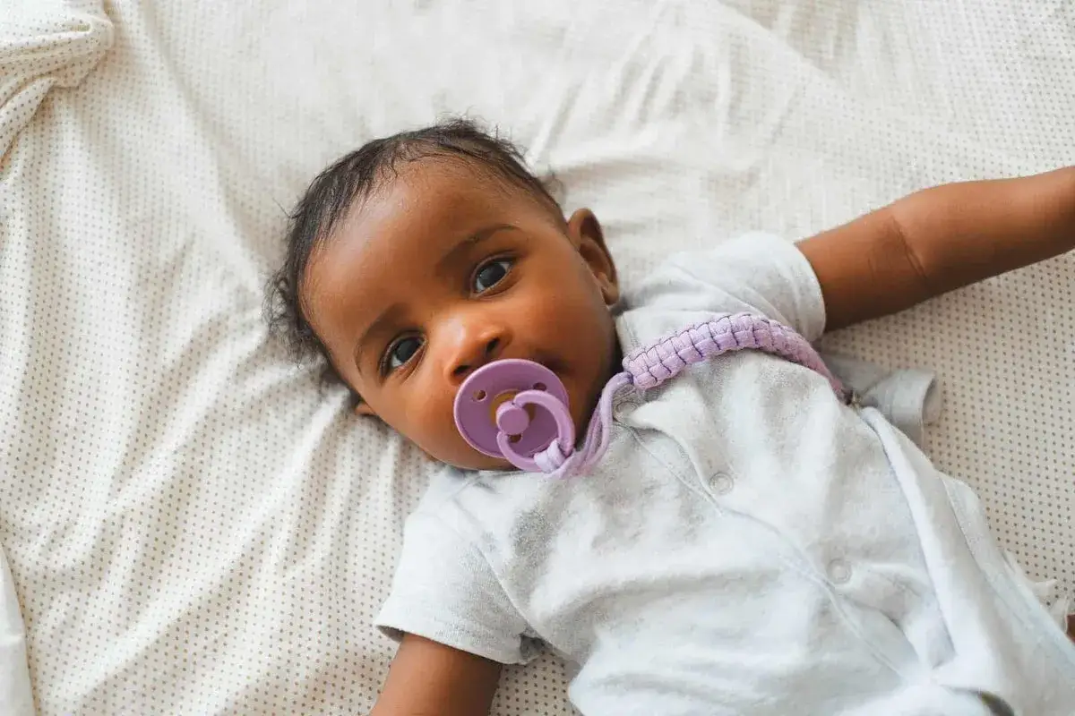 A baby with a binkie mouth lies on a patterned sheet, looking at the camera.