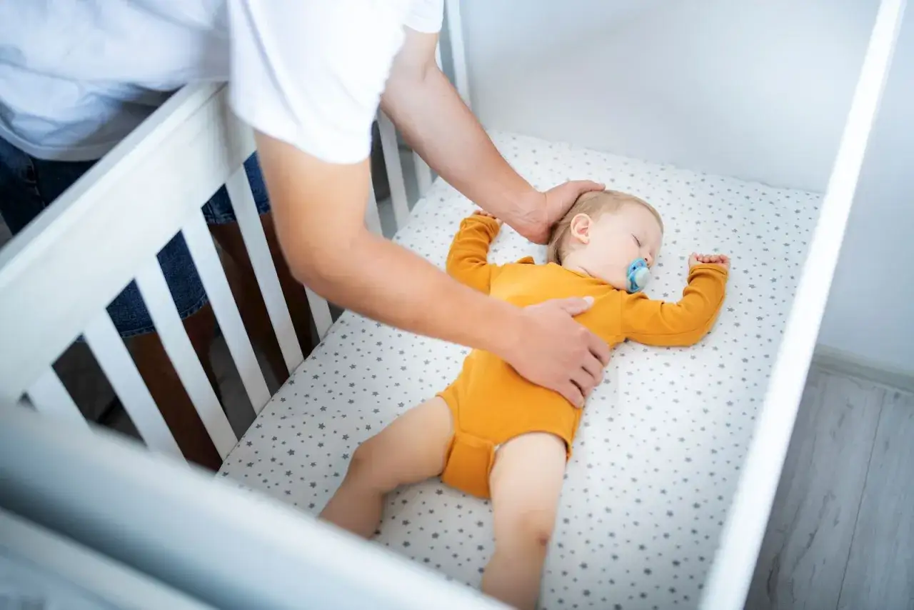 A father tucks his 8-month-old baby into a crib. The baby sleeps soundly with a pacifier, wearing an orange onesie.