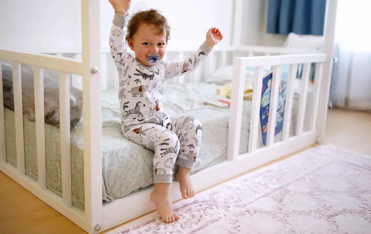A happy toddler in animal pajamas sits on a low bed with rails, perfect for what kind of bed should a 2 year old be in.