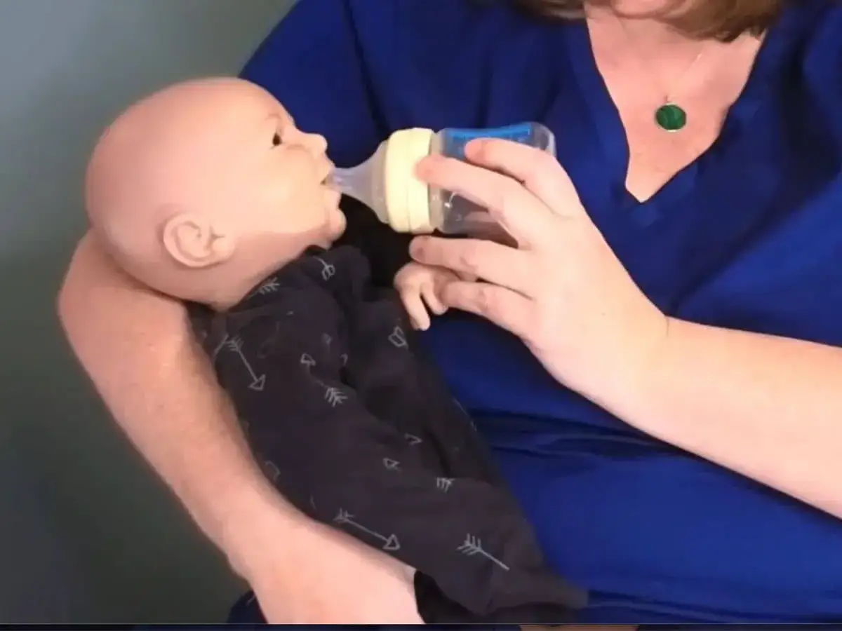 A person demonstrates paced feeding newborn with a bottle. The baby doll wears a dark onesie with arrows.