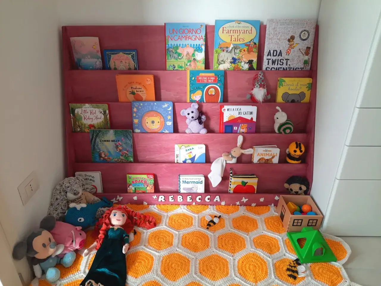 A DIY Montessori bookshelf filled with colorful children's books and plush toys. A handmade blanket with a honeycomb pattern lies on the floor.