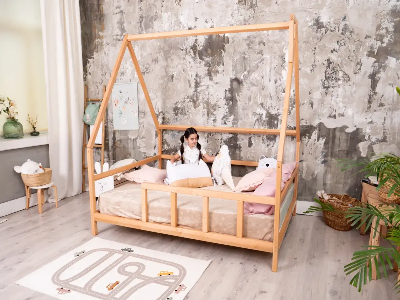 A young girl sits on a Montessori bed, surrounded by plush toys. The bed's natural wood frame and low profile are ideal for toddlers, and its dimensions are perfect for a child's room.