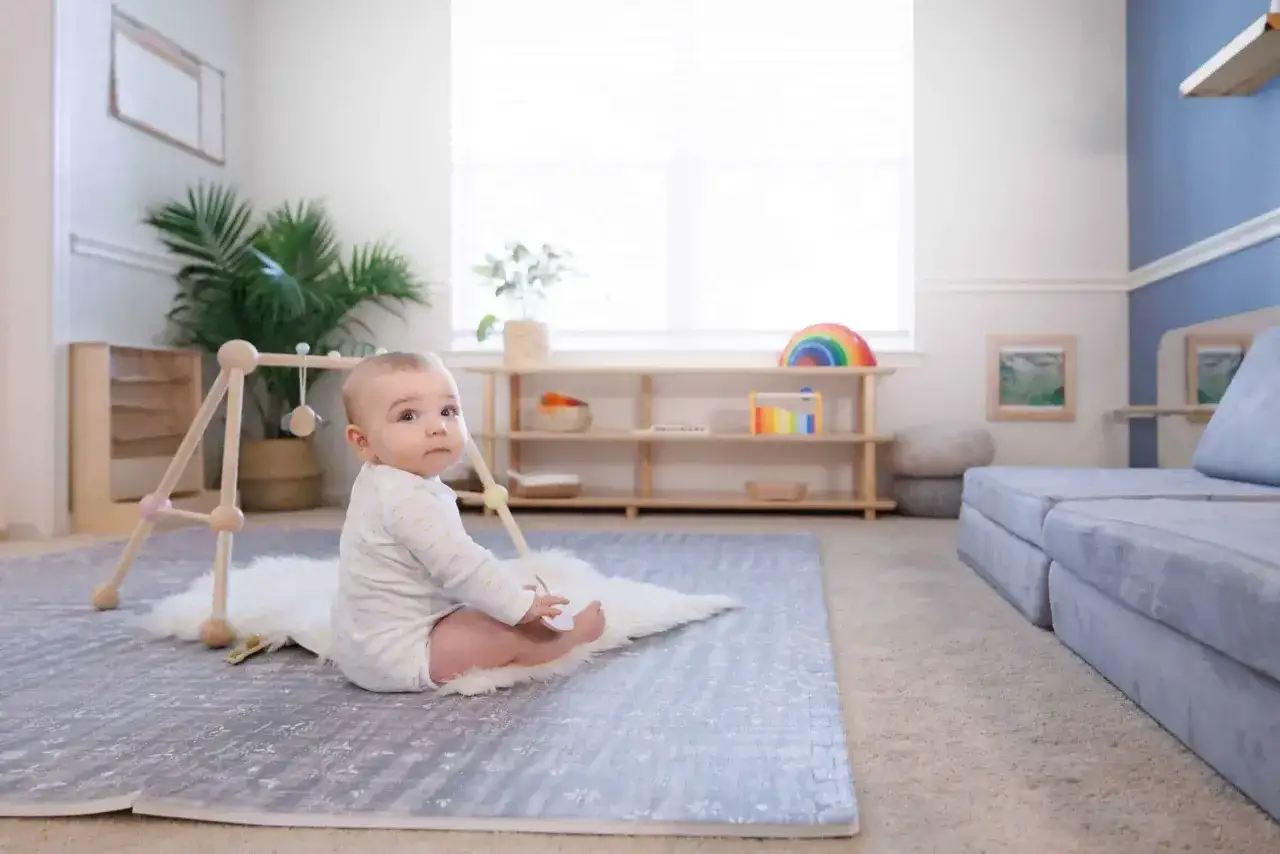 A baby sits on a fluffy rug in a room with a Montessori aesthetic, featuring wooden shelves, plants, and soft furnishings.