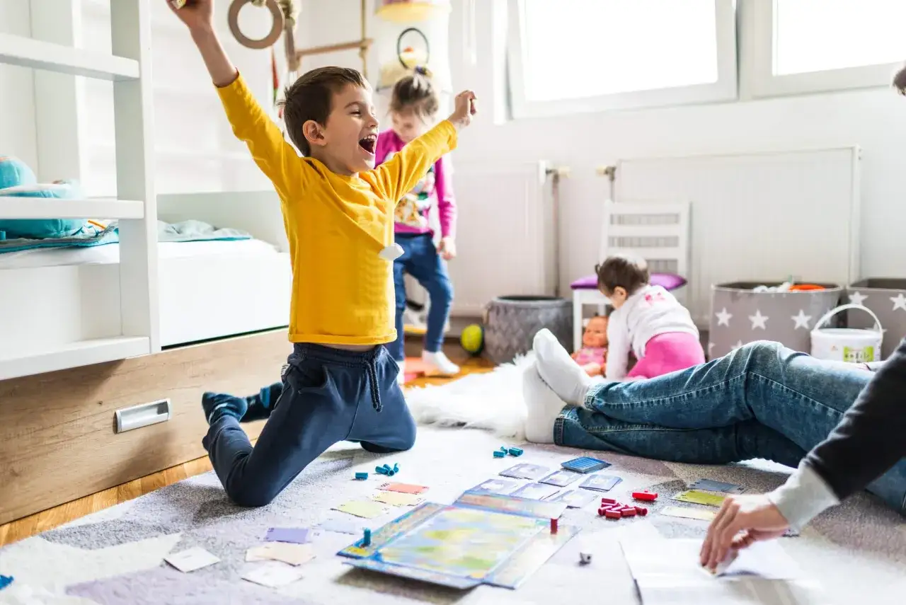 Excited boy celebrates winning a game, showing fun ideas for what to play indoors. Other children and an adult join the fun.