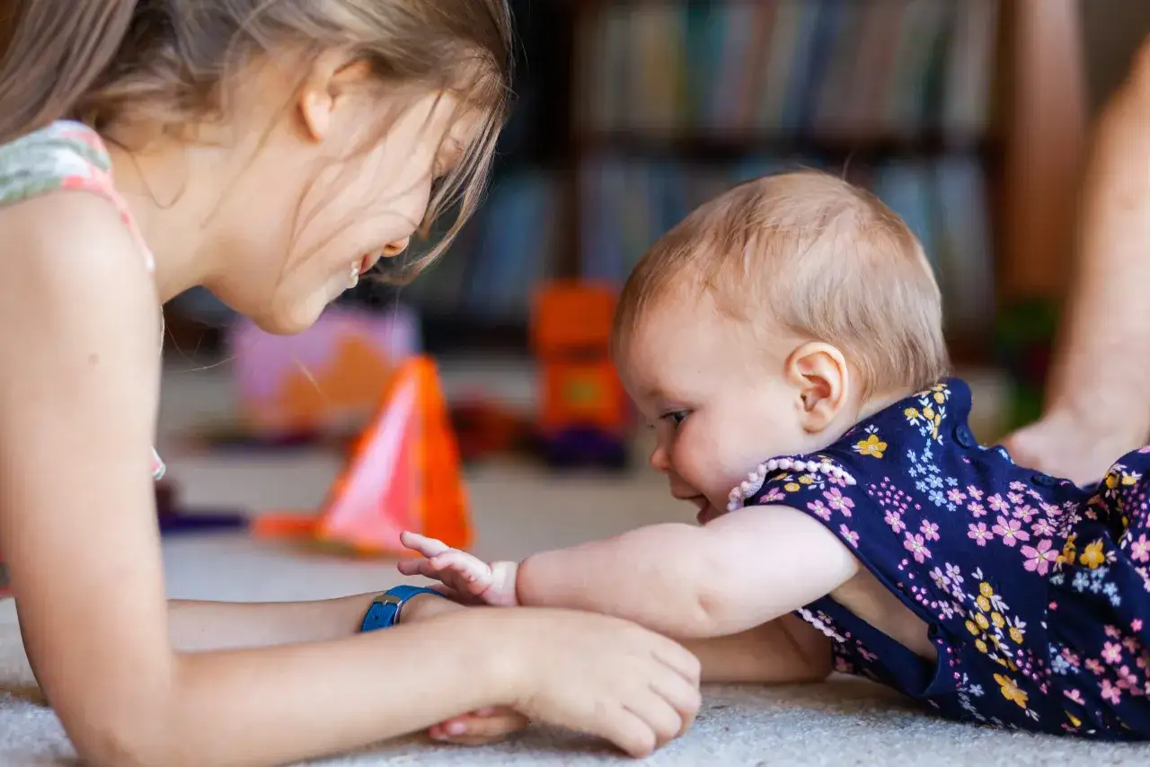 A young girl interacts with a baby, offering visual stimulation. The baby, lying on its stomach, reaches out to the girl.