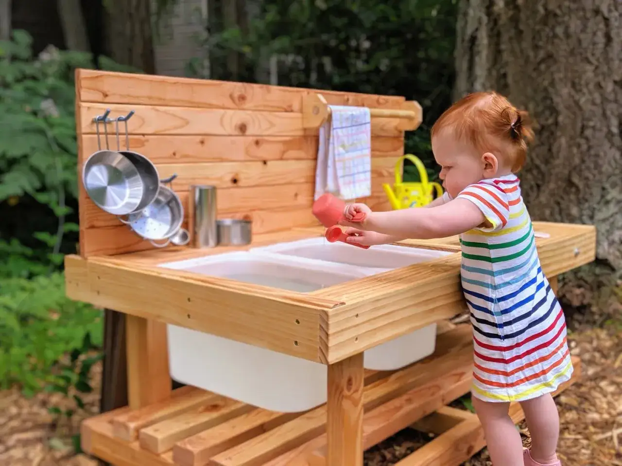 A toddler enjoys Montessori movement activities at a wooden mud kitchen, scooping water with red tools.