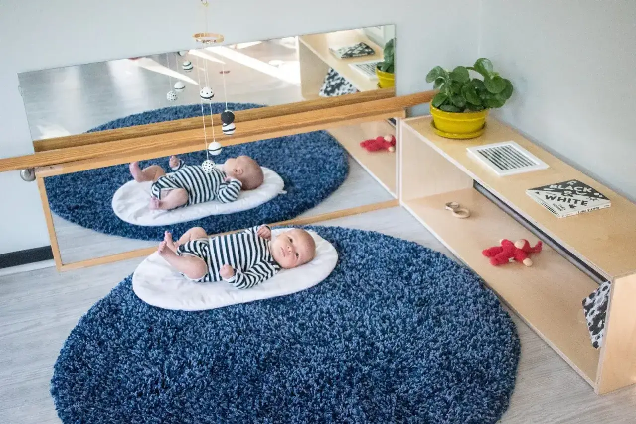 Two babies in striped onesies explore Montessori activities for babies, lying on blue rugs near a mirror and a shelf with toys and books.